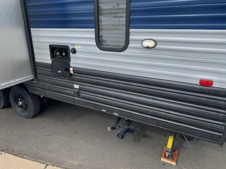 Side view of a parked blue and grey RV rental with an open door, showing external hookups and a stabilizing jack.. Cherokee Grey Wolf 27DBH 2021