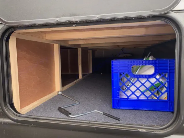 Pass-through RV rental storage bay with custom wood shelving, a blue crate, and a utility crank.. Cherokee Grey Wolf 27DBH 2021