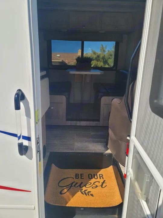 Welcoming RV rental interior with a 'Be Our Guest' mat, cozy dinette, and bright windows offering an outdoor view.. Forest River Sunseeker 2019