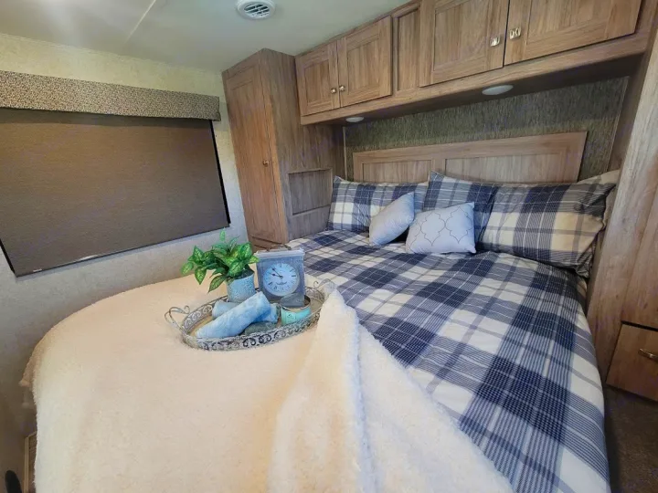 Cozy RV rental bedroom featuring blue and white plaid bedding, overhead storage cabinets, and a window with a roller shade.. Forest River Sunseeker 2019