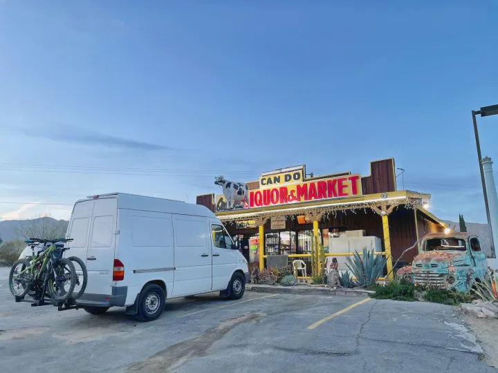 White Sprinter van, an RV rental option, carrying bikes, parked at Can Do Liquor & Market in a desert setting.. Mercedes Sprinter 2006