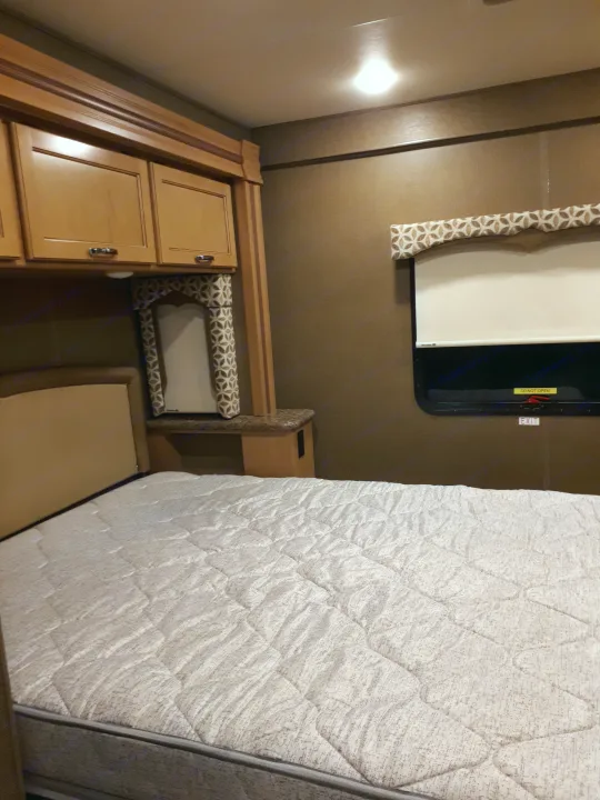 Inside an RV rental bedroom: a bed with a quilted mattress, wooden overhead cabinets, and a window with a shade and emergency exit sign.. Thor Four Winds 2017