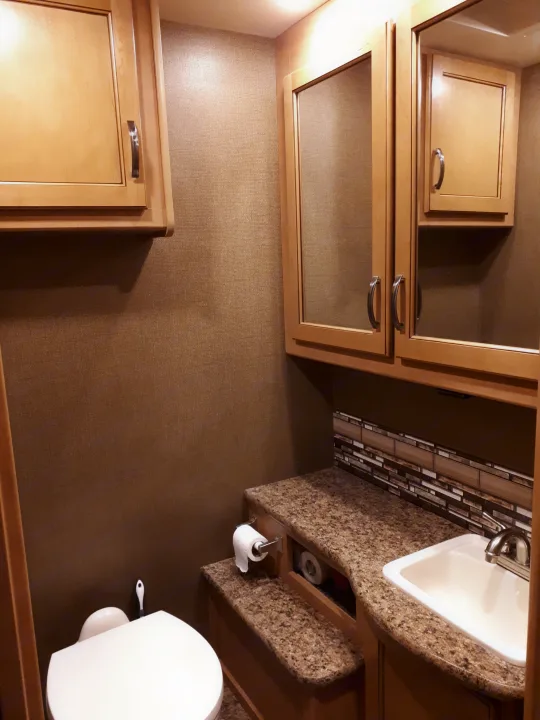 Cozy RV rental bathroom interior with light wood cabinets, speckled countertop, white sink, toilet, and textured brown walls.. Thor Four Winds 2017
