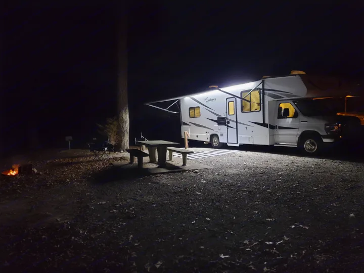 A bright RV rental illuminates a night campsite with a picnic table and a warm campfire glow. Perfect for outdoor camping.. Coachmen Freelander 2021