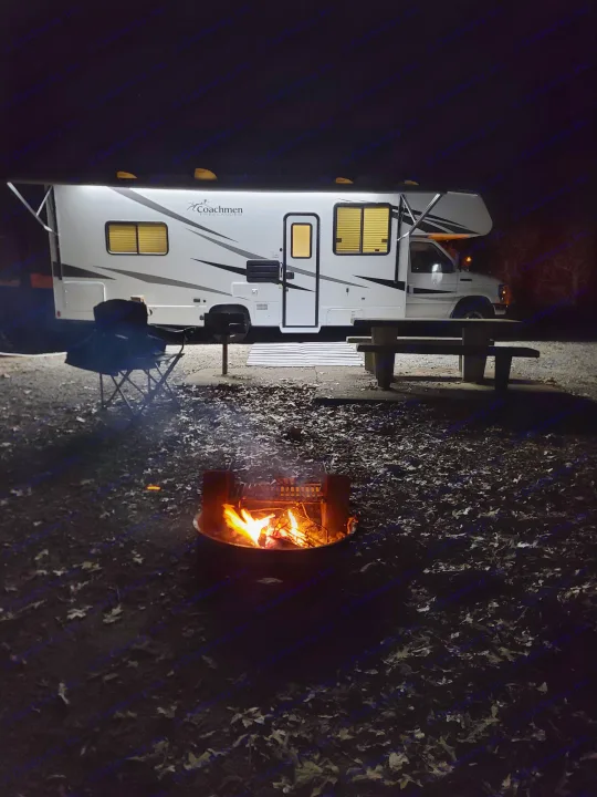 An RV rental campsite at night with a bright campfire, illuminated Coachmen RV, and picnic area. Perfect for outdoor adventures.. Coachmen Freelander 2021