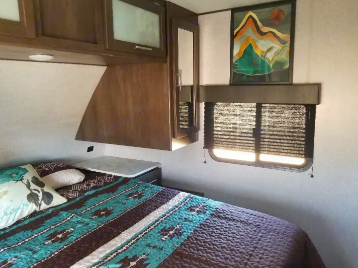 RV rental bedroom interior: a bed with patterned comforter, overhead wood cabinets, and a window with black blinds.. Dutchmen Coleman 2021
