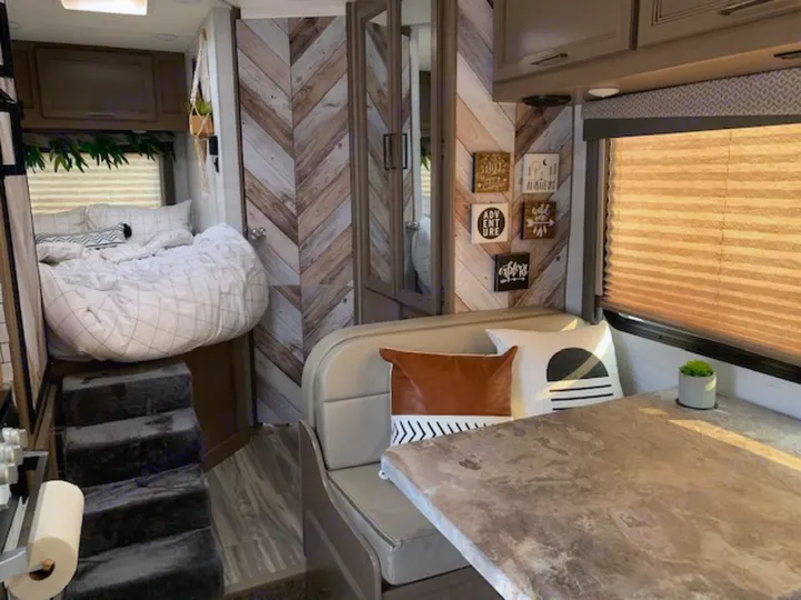 Cozy RV rental interior with elevated bed, steps, and inviting dinette featuring decorative pillows & accents.. Thor Four Winds 2021