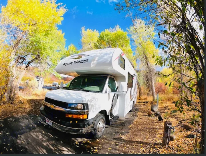 Scenic RV rental in autumn. A white camper sits on a leafy path, surrounded by vibrant yellow trees under a clear blue sky.. Thor Four Winds 2021