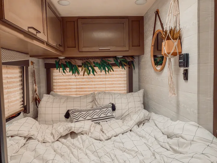 Cozy RV rental bedroom: white grid bedding, wooden cabinets, window blinds, and stylish decor including a hanging plant and mirror.. Thor Four Winds 2021