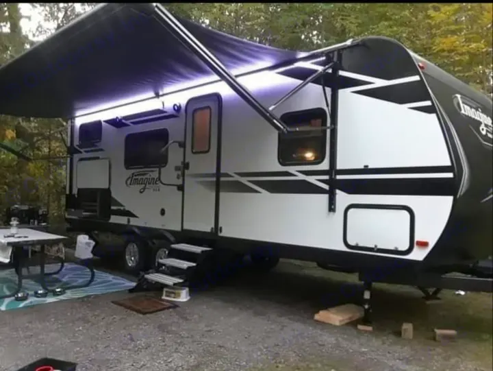 This Imagine travel trailer RV rental is set up at a scenic campsite for outdoor living, featuring an extended awning with lights.. Grand Design Imagine 23 BHE 2021