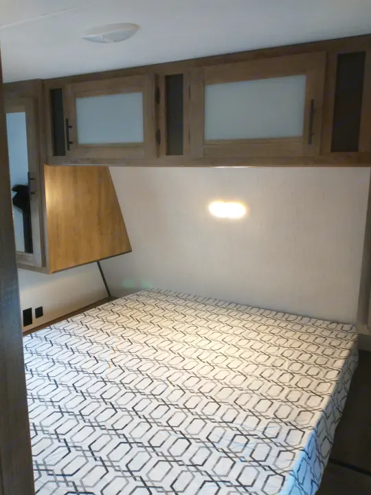 RV rental bedroom with a geometric bedspread, overhead storage cabinets, and a wall-mounted light.. Heartland Pioneer 2021