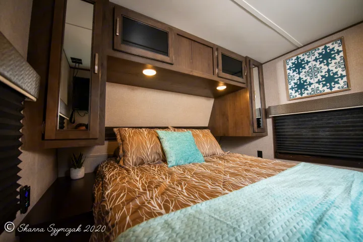 Inside an RV rental bedroom: cozy bed with patterned comforter, overhead cabinets, warm lights, and decorative art.. Jayco Jay Flight 2020
