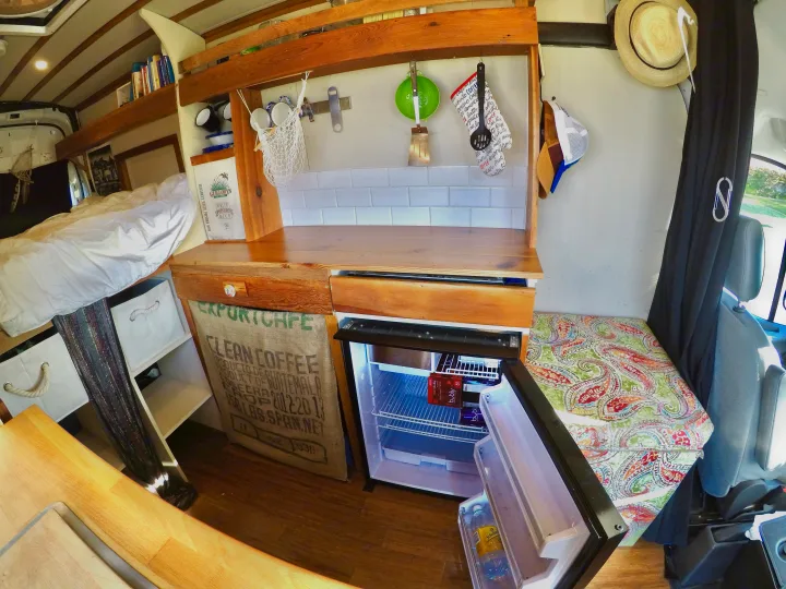 An RV rental interior showing a compact kitchen with wood counters, bed, and opened fridge with drinks.. Ford Transit 2017