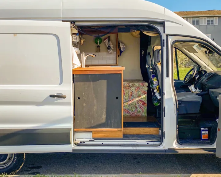 Inside this white RV rental van, a custom conversion reveals a compact kitchen with a sink, plus a colorful bench and front cab.. Ford Transit 2017