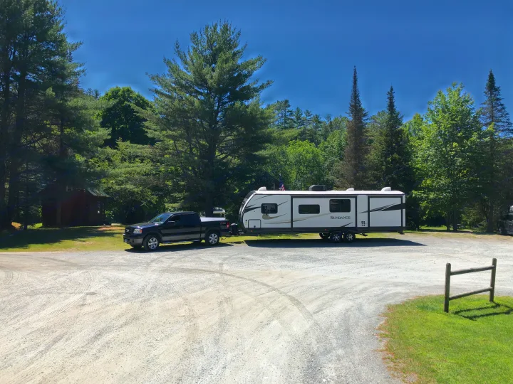 Enjoy an RV rental getaway! A pickup truck tows a large white travel trailer on a gravel campsite among tall trees and a cabin.. Heartland Sundance 2019