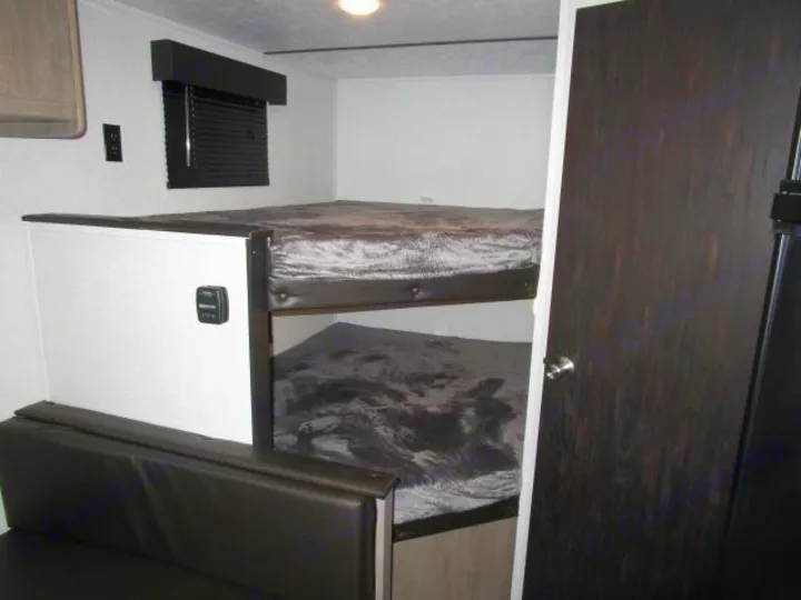 RV rental bunkhouse features dark bunk beds over a couch, with a window and a dark wood-grain door for a cozy space.. Keystone Hideout 2021