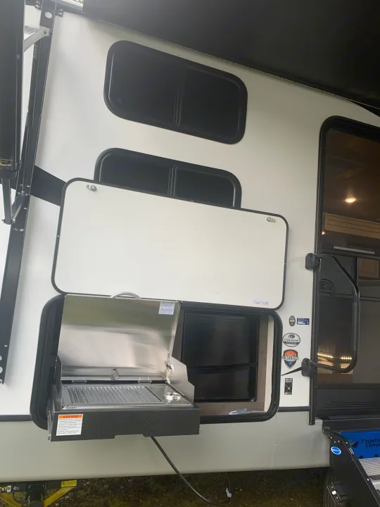 An RV rental showing its outdoor kitchen: a grill and fridge are revealed by an open hatch, perfect for camping trips.. Keystone Cougar Half-Ton 2021
