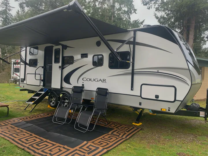 Modern white 'Cougar' RV rental parked on grass with extended awning, chairs, and patio mat.. Keystone Cougar Half-Ton 2021