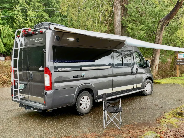 This Winnebago Travato RV rental is parked with its awning extended at a campsite, perfect for outdoor relaxation.. Winnebago Travato 2018