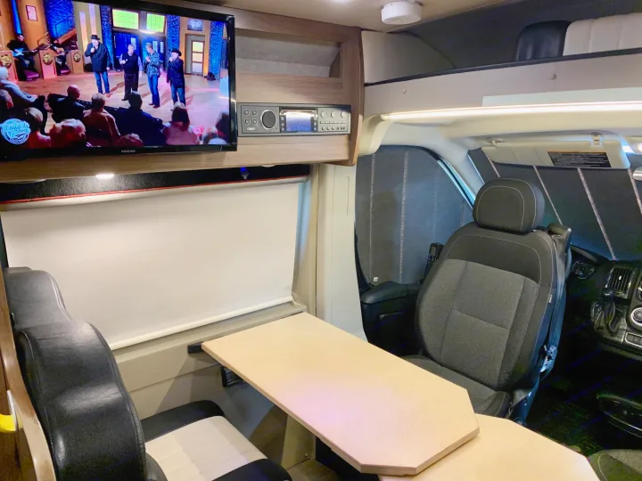 Inside an RV rental, a TV plays in the lounge area with a dining table and front driver's seat visible.. Winnebago Travato 2018
