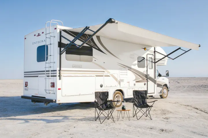 White Entegra Odyssey RV rental with extended awning and camping chairs on a flat, sandy landscape near water.. Entegra Coach Odyssey 25R 2019
