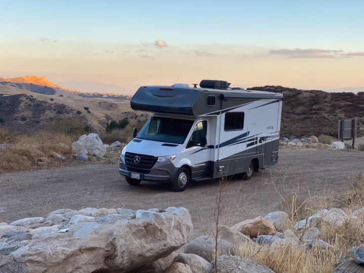 A white and gray Vita RV rental parked on a scenic dirt road, with hills and a distant town in the golden hour light.. Winnebago Vita 2020