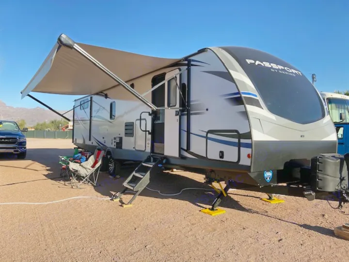 Sunny day at the campsite with a modern RV rental, awning extended, providing shade for outdoor relaxation and travel fun.. Keystone RV Passport 2021