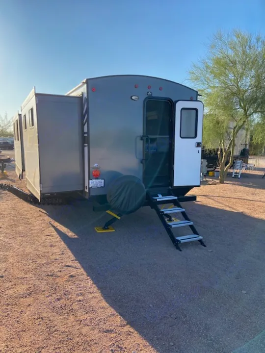 Grey RV rental with extended slide-out, rear ladder, spare tire, and yellow jacks, parked on gravel.. Keystone RV Passport 2021