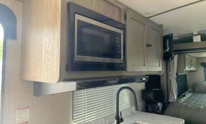 Inside this RV rental, a bright kitchen features an over-counter microwave, sink, and wood-grain cabinets. A cozy bed is seen beyond.. Keystone Outback Ultra-Lite 2021