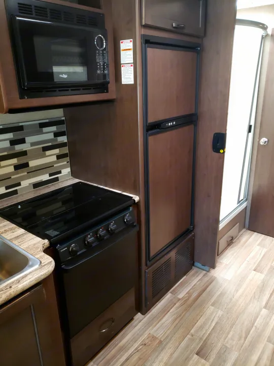 Compact RV rental kitchen features built-in microwave, fridge, and stove with modern dark wood cabinets and light flooring.. Thor Motor Coach 30.3 2019