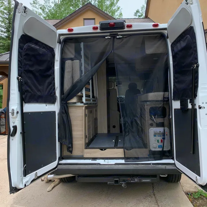 This RV rental camper van's open rear doors show a mesh screen and custom interior with a bed/kitchen. Dog paws peek underneath.. Winnebago Solis 2021