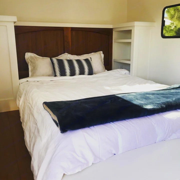 Experience comfort! This RV rental bedroom features a cozy bed with white linens, a wooden headboard, and built-in storage by a window.. Custom Camper Trailer 2020