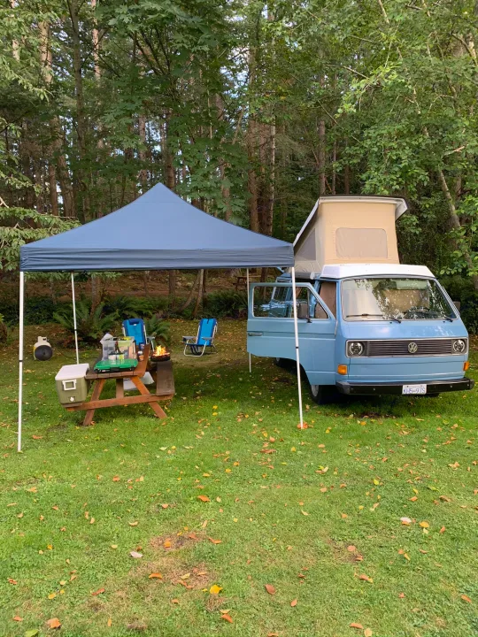 Classic blue VW camper van with pop-top, at a grassy forest campsite with a canopy, picnic table, and fire. Perfect RV rental for camping.. Volkswagen Westfalia 1981