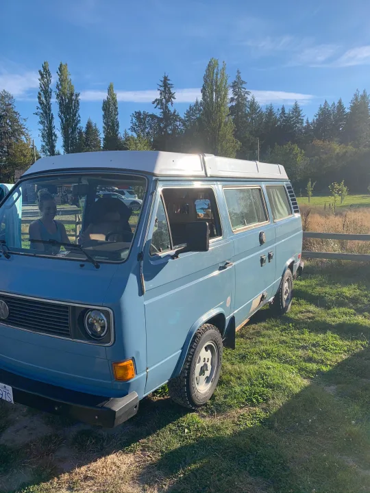 A blue VW Vanagon camper van is parked on grass with a smiling driver. Ideal rv rental for outdoor adventures.. Volkswagen Westfalia 1981
