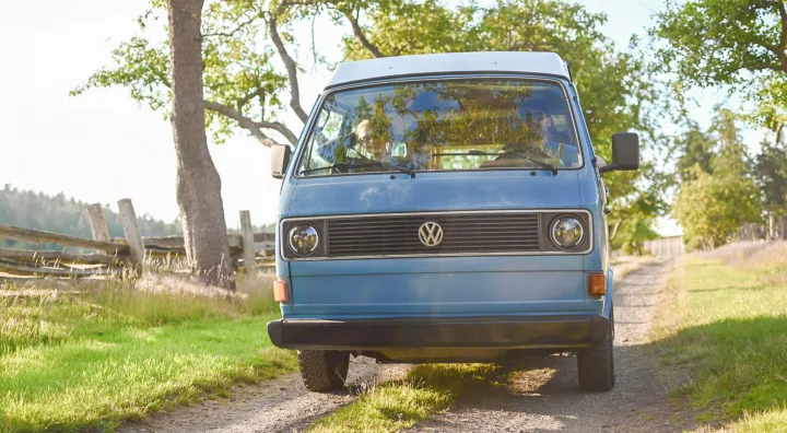 Light blue VW camper van, an ideal RV rental, on a dirt road. Trees and a wooden fence frame the scene.. Volkswagen Westfalia 1981