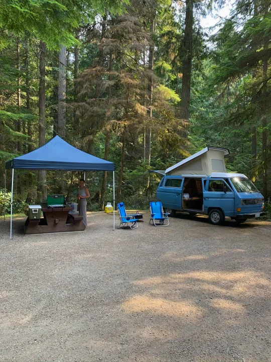 A blue camper van, perfect as an RV rental, is set up at a serene forest campsite with a canopy and picnic area.. Volkswagen Westfalia 1981