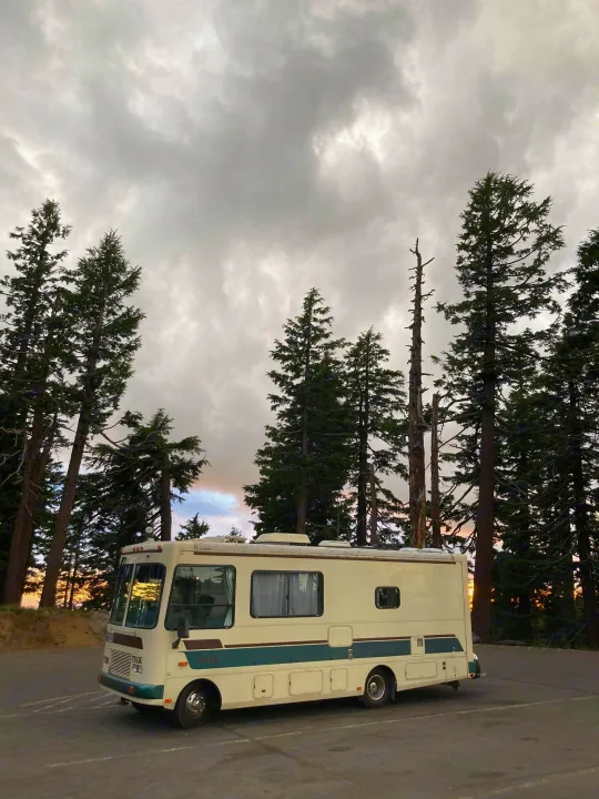 An RV rental parked under a cloudy sky among tall pines, with sunset glow through the trees.. Safari Trek 2610 1994