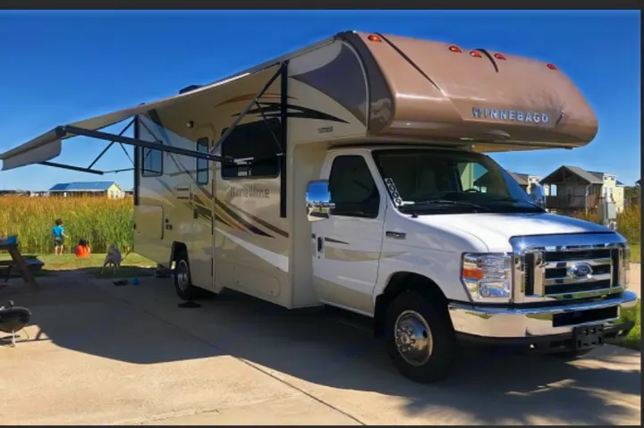 White & beige Winnebago RV rental, awning extended, parked at a sunny site. A child & dog are in the background.. Winnebago Minnie Winnie 2019