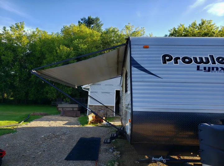 A Prowler Lynx RV rental with a deployed awning, steps down, parked on gravel near a partial build and lush trees.. Heartland Prowler Lynx 2014