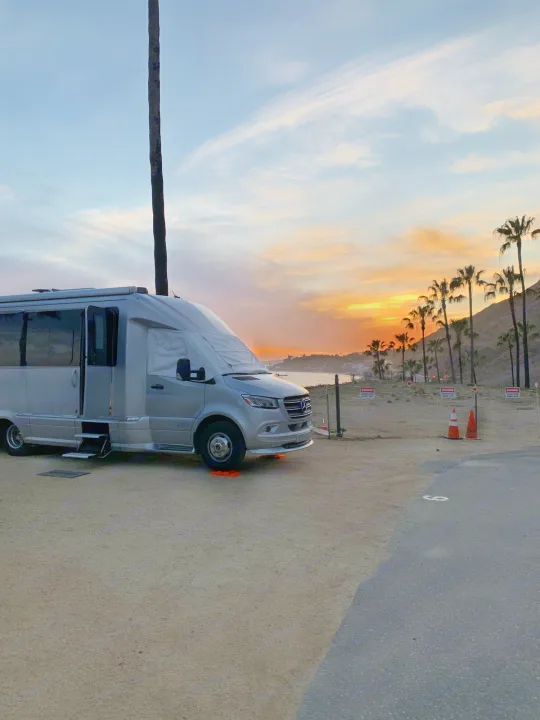 An rv rental overlooks a beautiful sunset bay, with a silver RV on sand, silhouetted palm trees, and a vibrant sky.. Airstream Atlas 2021