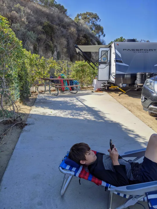 A boy rests in a lounge chair, looking at his phone, while an RV rental is parked nearby at a sunny campsite.. Other Other 2021