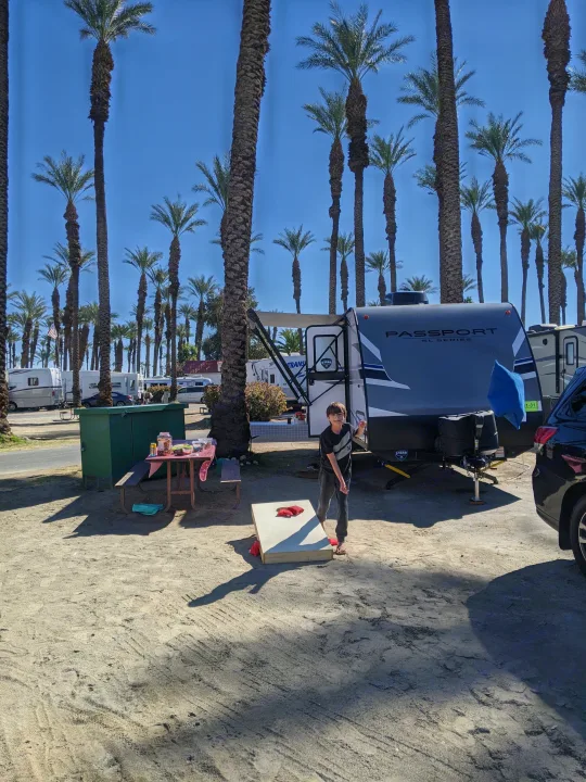Sunny RV park scene: boy plays cornhole by a Passport SL Series rv rental, surrounded by tall palm trees.. Other Other 2021