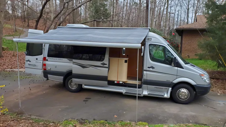 Silver Pleasure-Way Sprinter **rv rental** with extended awning & open door, parked on a driveway near trees.. Pleasure Way Plateau Ts 2013