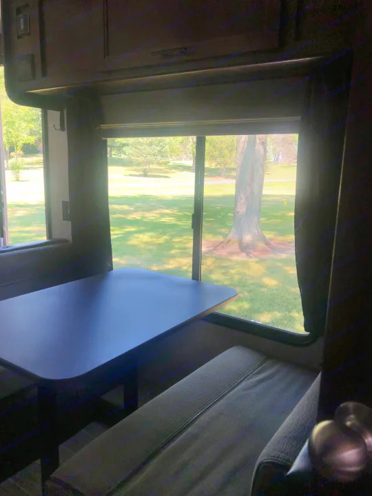 Inside an RV rental: a dark dinette table and seating, with a large window view of a sunny, grassy park.. Forest River Cherokee Grey Wolf 22MKSE 2020