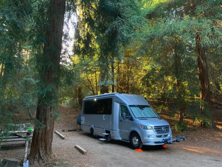 Silver RV rental settled at campsite 104 among towering trees. Ideal for a serene forest getaway, with a picnic area and bike rack.. Airstream Atlas 2021
