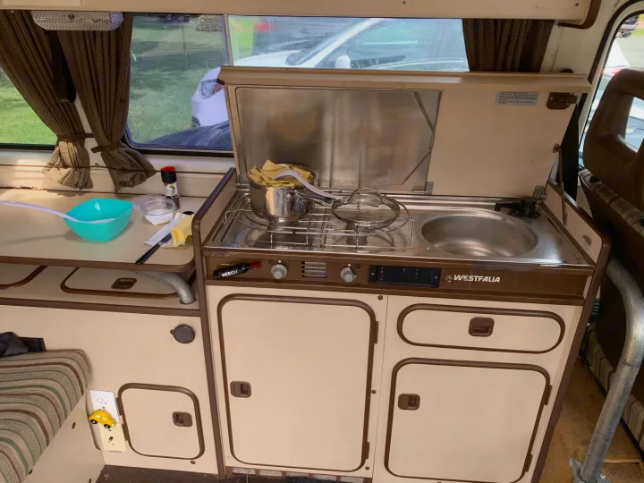 Cooking a meal in a cozy Westfalia RV rental, featuring a compact kitchenette with stove, sink, and cabinets.. Volkswagen T3 Westfalia 1984