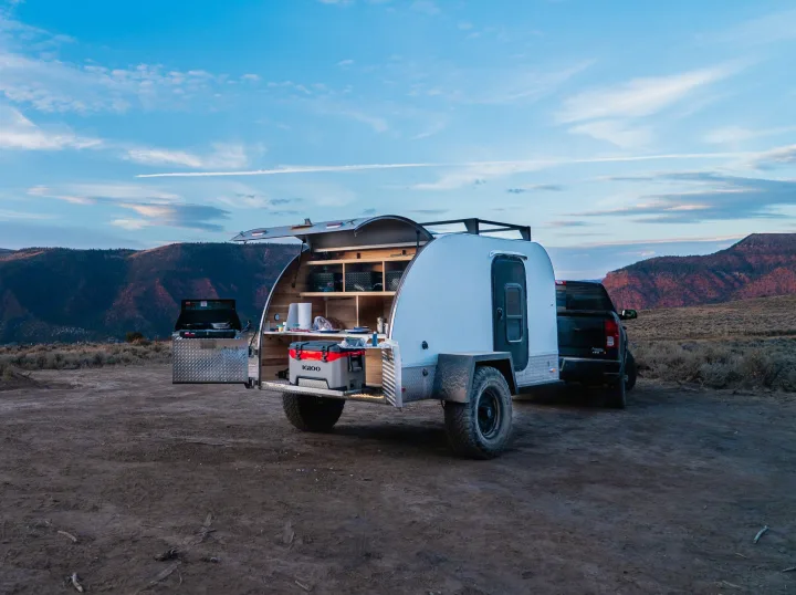 Outdoor rv rental adventure: A white teardrop camper with an open rear kitchen, including an Igloo cooler, hitched to a truck at dusk.. PINNACLE Pinnacle Summit 2020