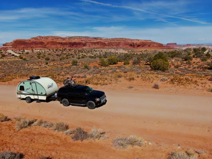 Black SUV with bike towing an RV rental teardrop trailer on a desert dirt road, red rock formations under blue sky.. Other Other 2016