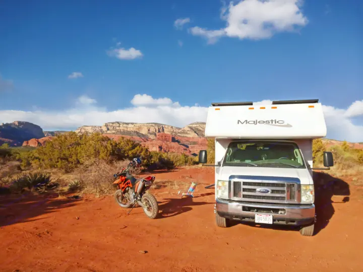 Adventure awaits! This white RV rental and dirt bike are parked on red dirt, surrounded by stunning red rock formations in a sunny, arid landscape.. Four Winds Majestic 2009