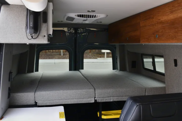 View inside an RV rental's camper van, showing a grey rear bed, wooden overhead cabinets, AC unit, and side windows.. Storyteller Overland Storyteller Overland Class B 2021